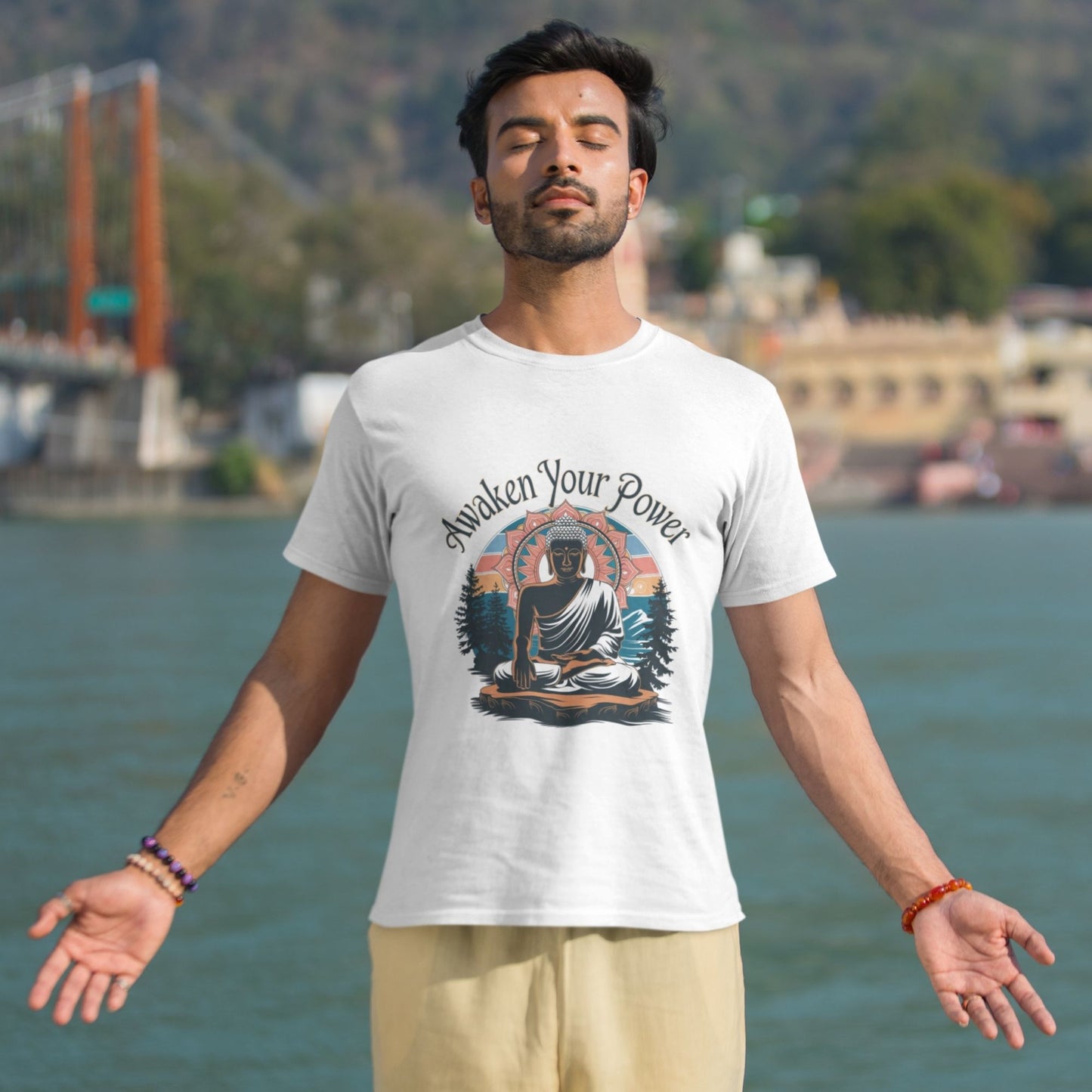 A man in a white 'Spiritual Awakening Tee' with a Buddha design meditates outdoors with eyes closed, arms outstretched