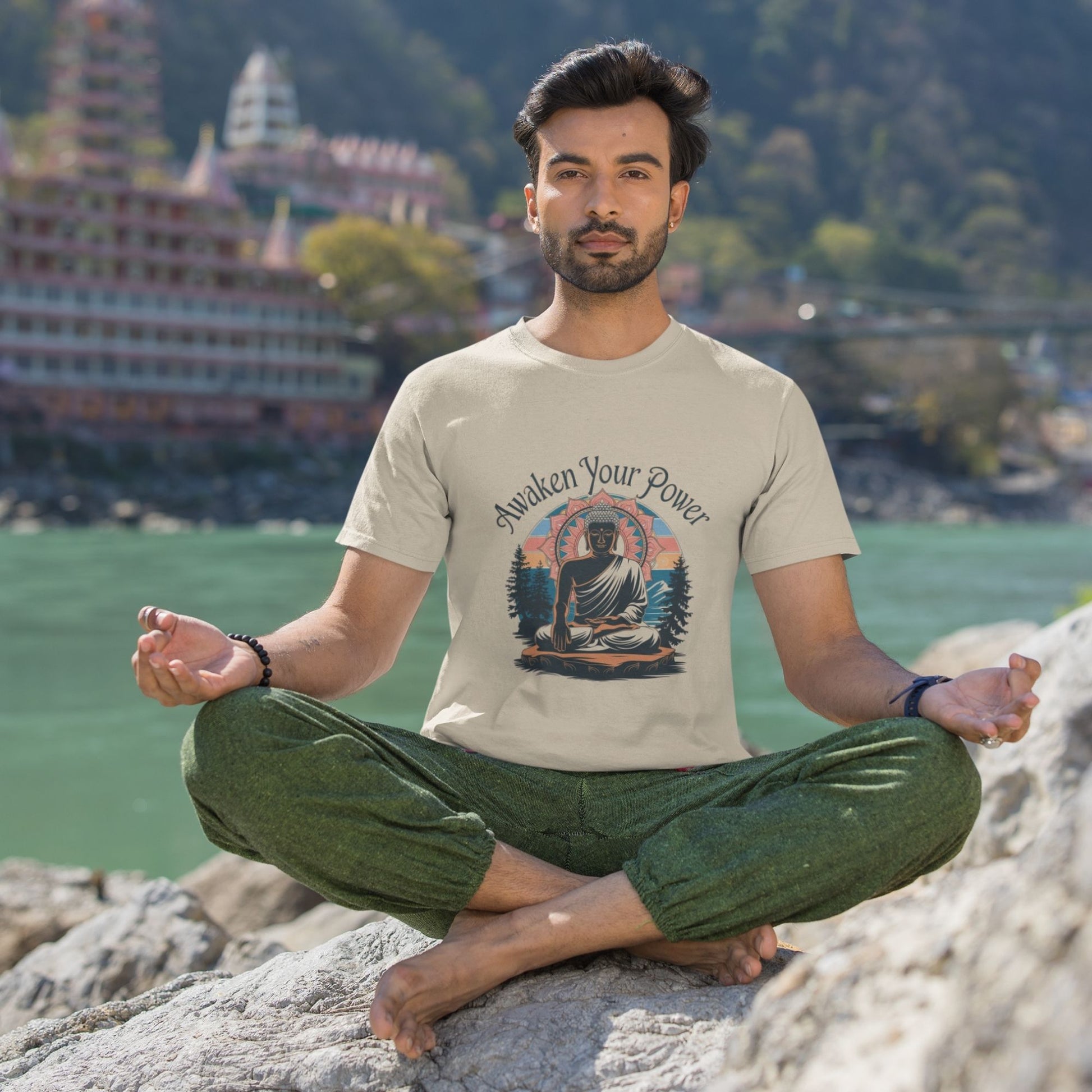 Man in a light-colored zen graphic shirt in lotus pose on rocks by water with buildings in background