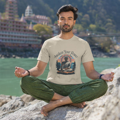 Man in a light-colored zen graphic shirt in lotus pose on rocks by water with buildings in background
