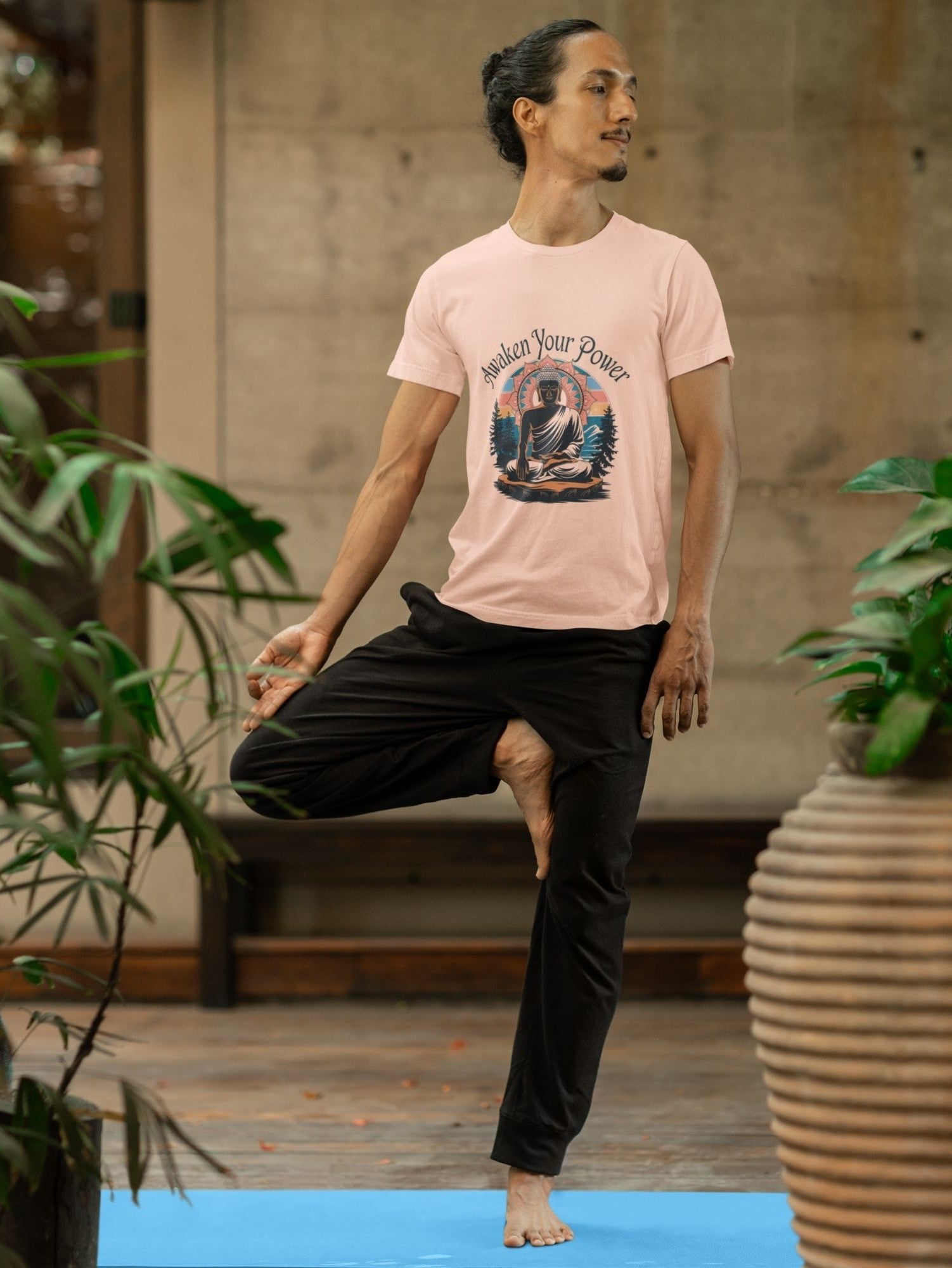 Man in peach Buddha meditation t-shirt color doing tree pose on a blue yoga mat, surrounded by plants.
