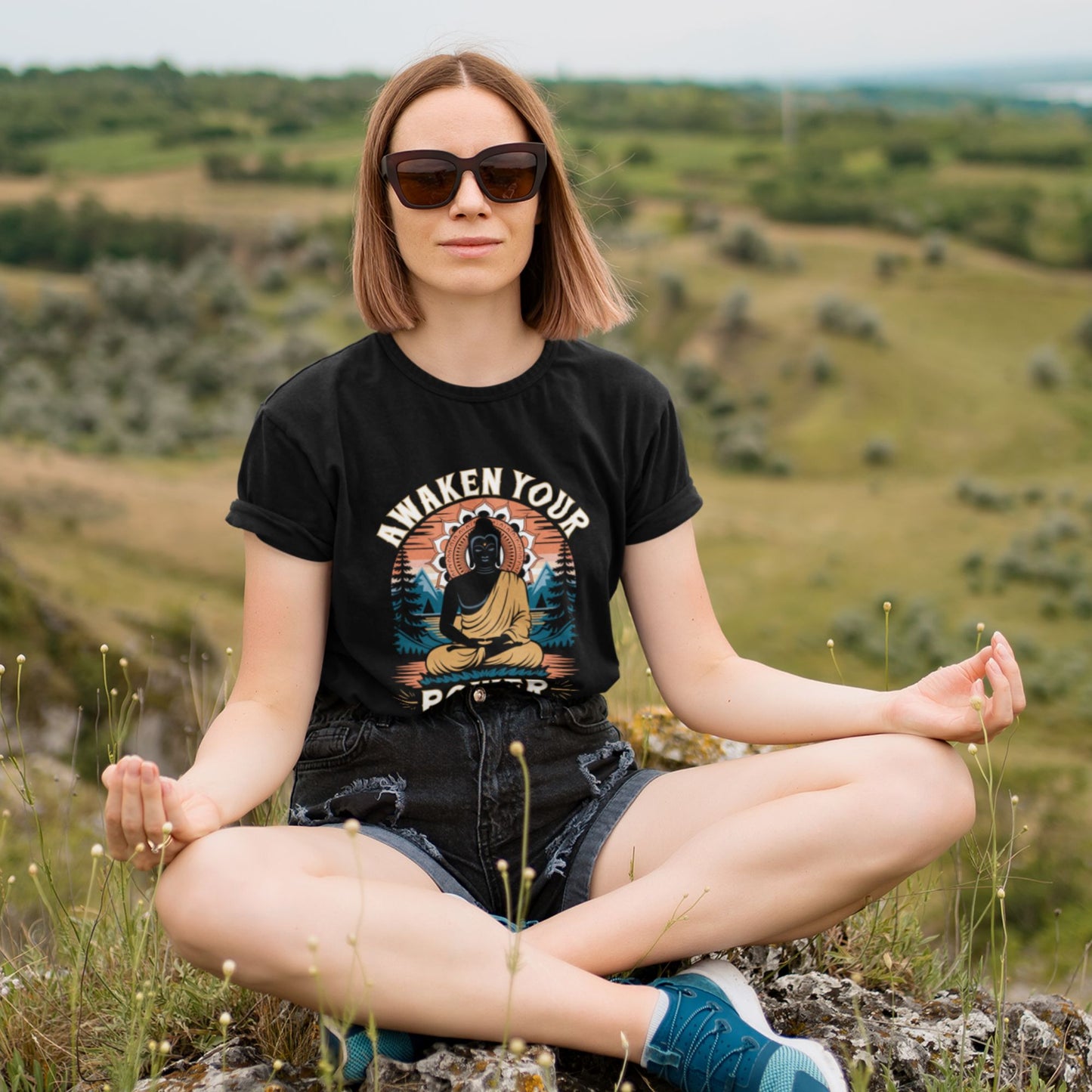 Woman sitting outdoors in a field wearing a black t-shirt with a graphic design.