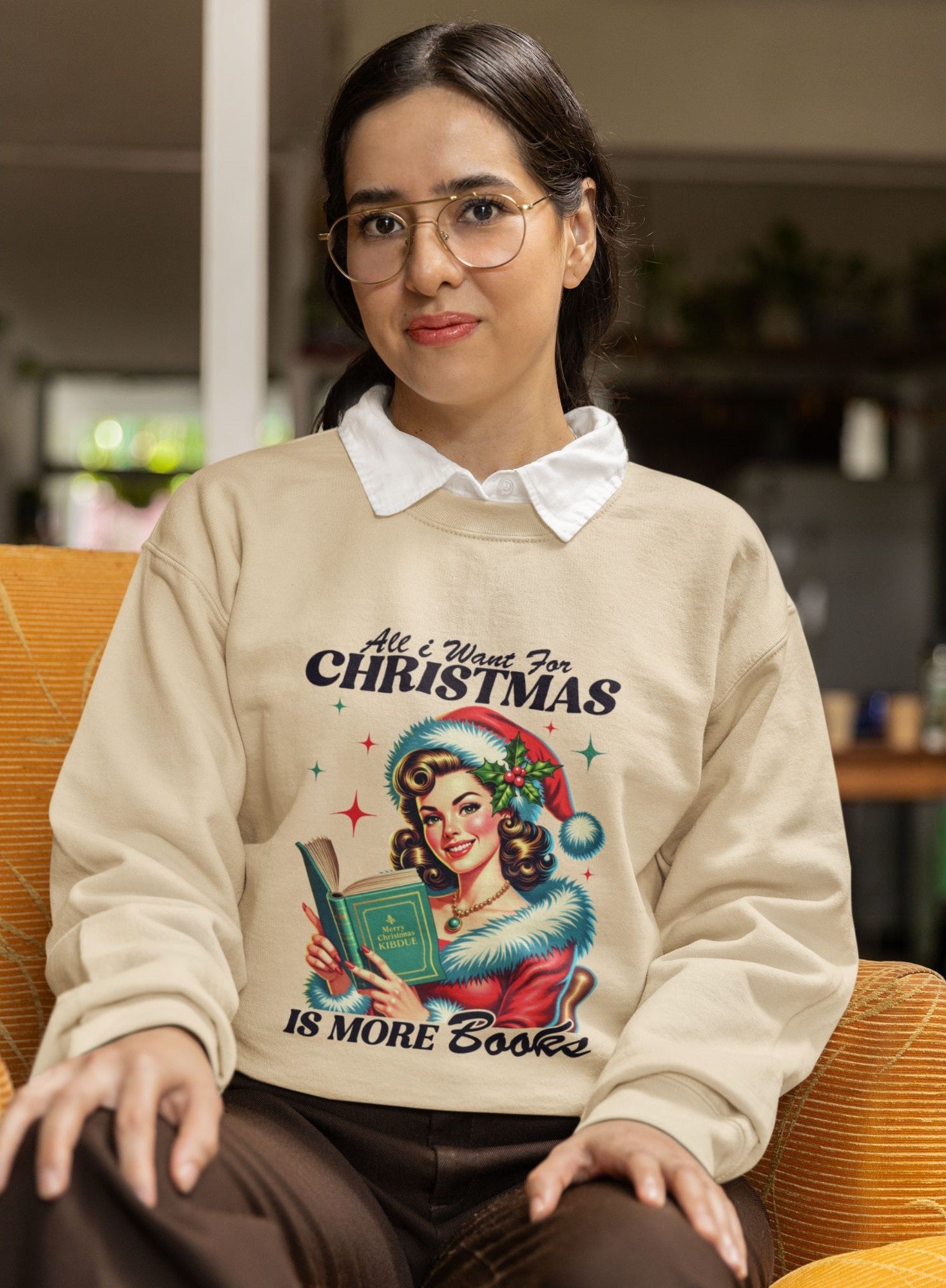 Woman-wearing-a-beige-Christmas-themed-sweatshirt-reading-All-I-Want-for-Christmas-is-More-Books-seated-on-a-mustard-armchair