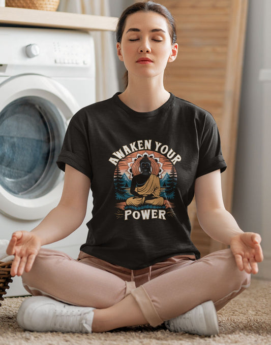 A woman sits in a meditative pose on a bed, wearing a black Zen Buddha tee that says "AWAKEN YOUR POWER."
