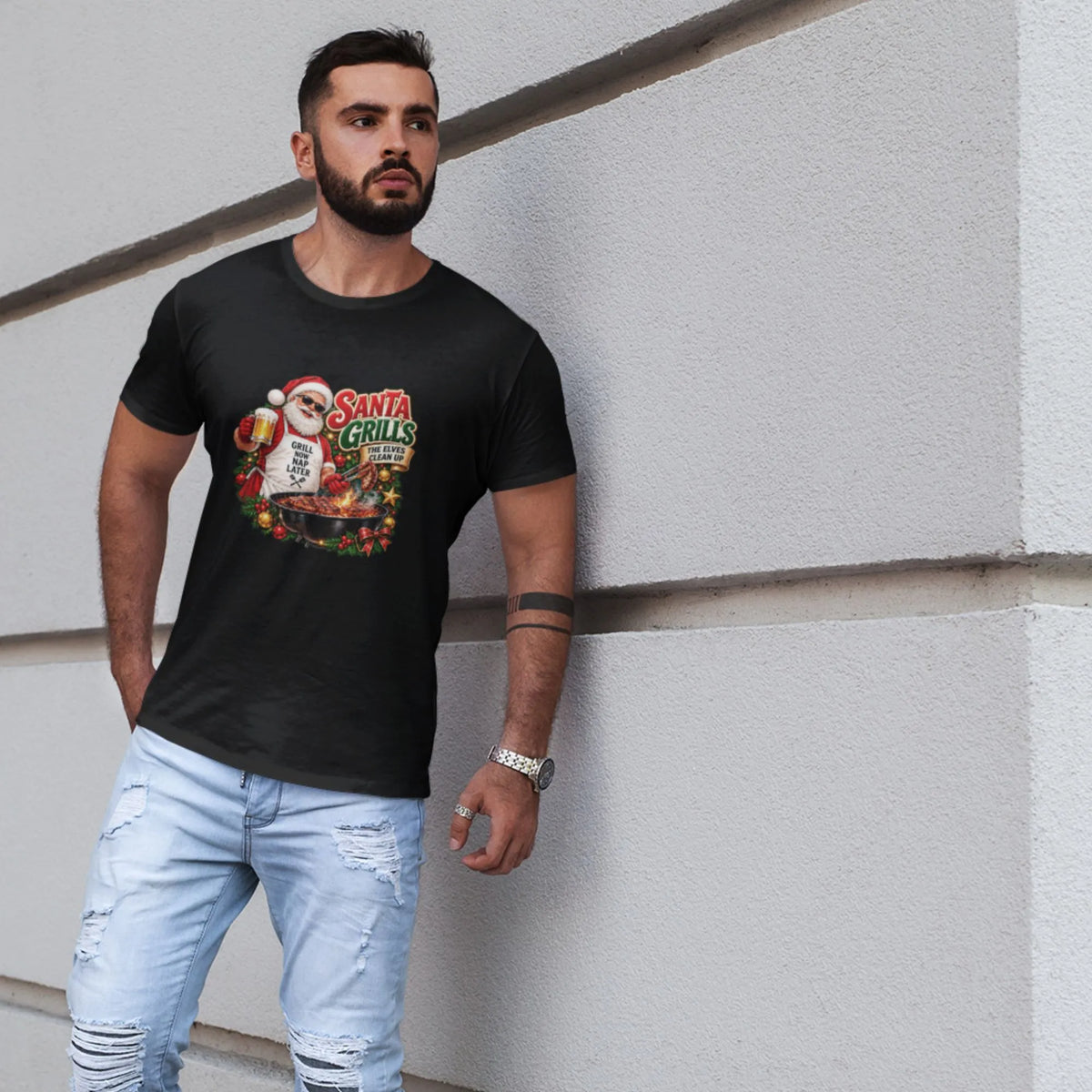A man wearing a black graphic t-shirt with a Santa Claus and 'Santa Grills' design, paired with light-wash distressed jeans, leaning against a light gray textured wall.