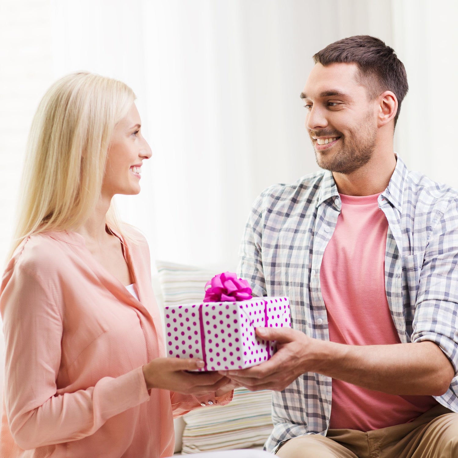 A smiling man in a plaid shirt is giving a polka-dotted gift box with a pink bow to a smiling woman.