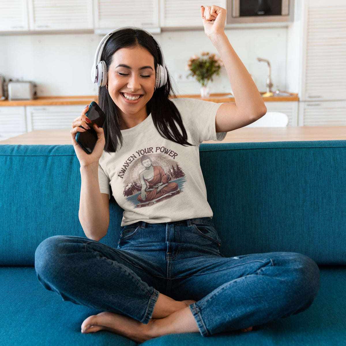 A happy woman wearing an inner peace t-shirt with a Buddha graphic and the text "AWAKEN YOUR POWER" is sitting on a blue couch at home, enjoying music with headphones and holding a smartphone.