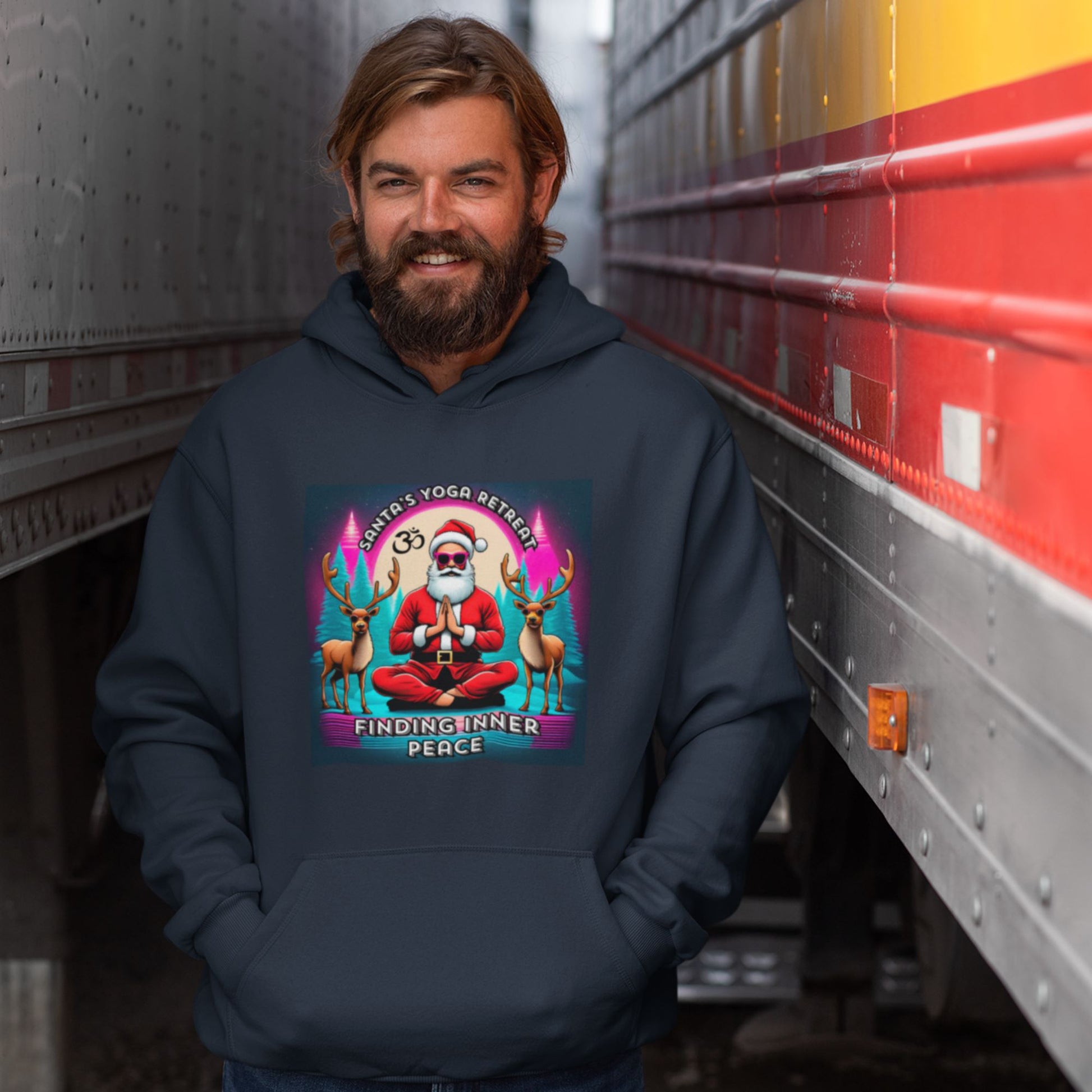 A smiling man wearing a dark-colored graphic hoodie featuring a cartoon image of Santa Claus meditating with reindeer, standing between two large trucks