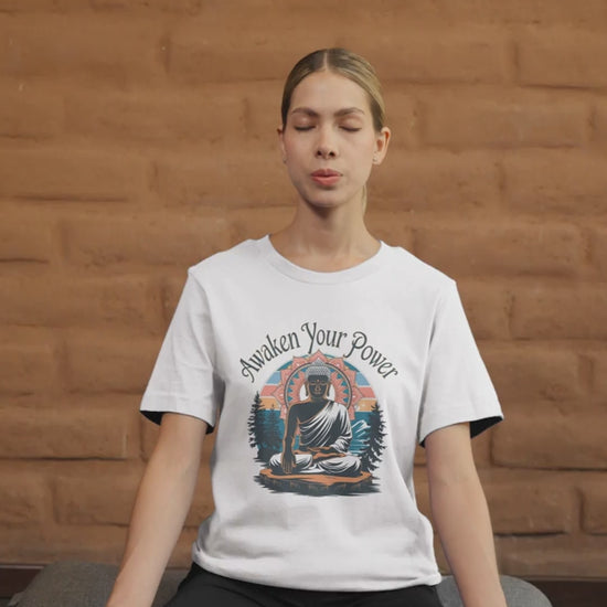 A woman meditates cross-legged on a mat, eyes closed and hands resting on her knees. She wears a white "Zen Power" t-shirt featuring an image of Buddha