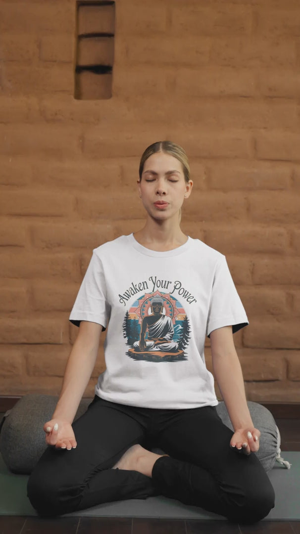 A woman meditates cross-legged on a mat, eyes closed and hands resting on her knees. She wears a white "Zen Power" t-shirt featuring an image of Buddha