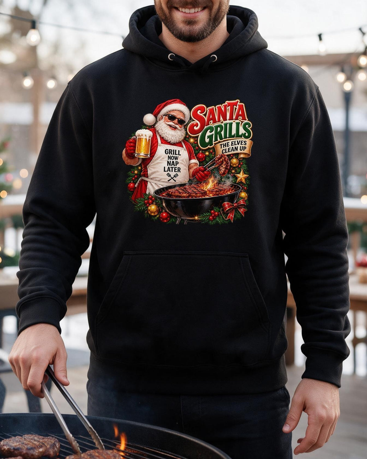 Man wearing a black Santa grilling Christmas hoodie, using tongs to cook meat on a barbecue with the chest print visible