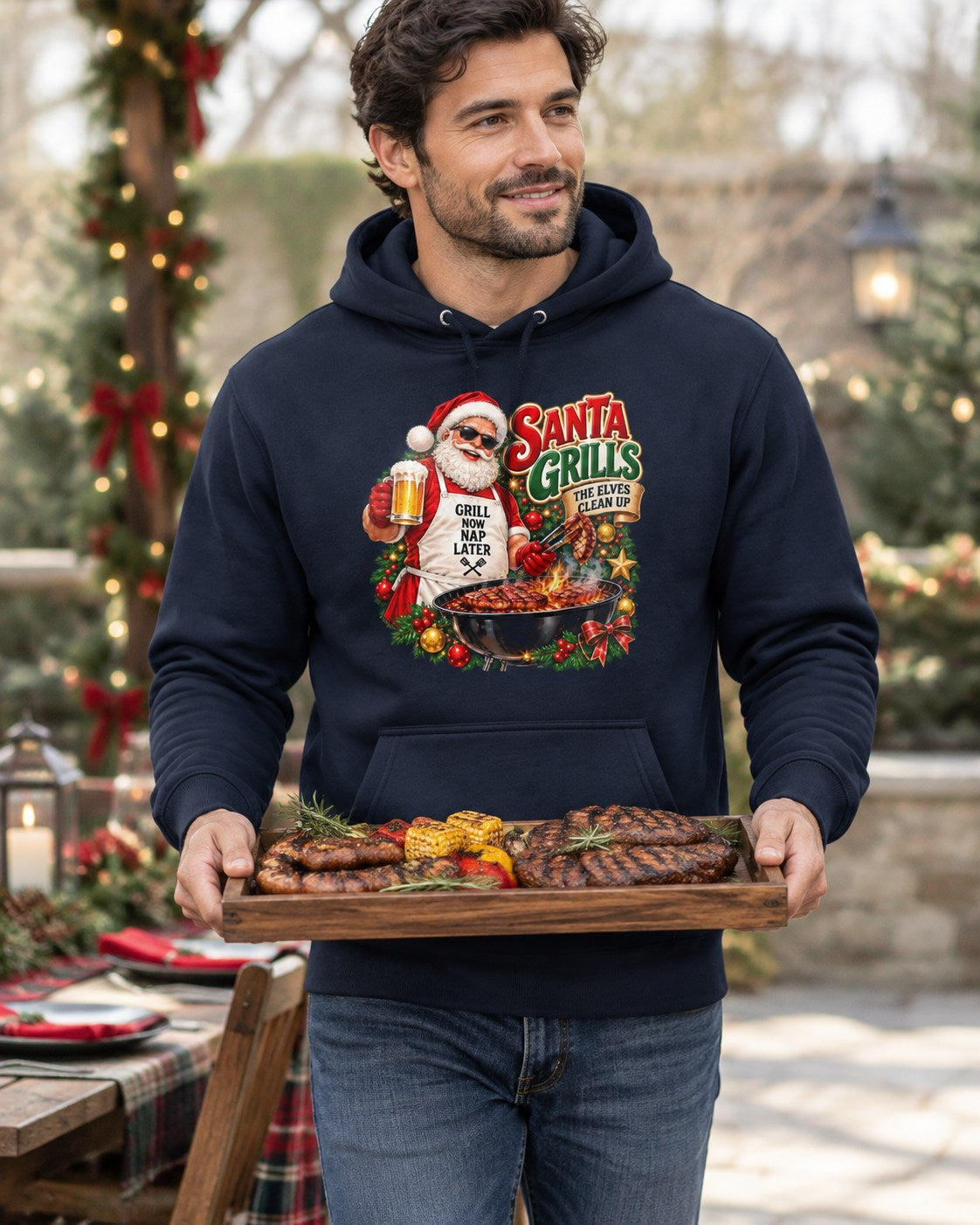 Man wearing a navy Santa grilling Christmas hoodie, carrying a tray of grilled meat and corn beside a festive table