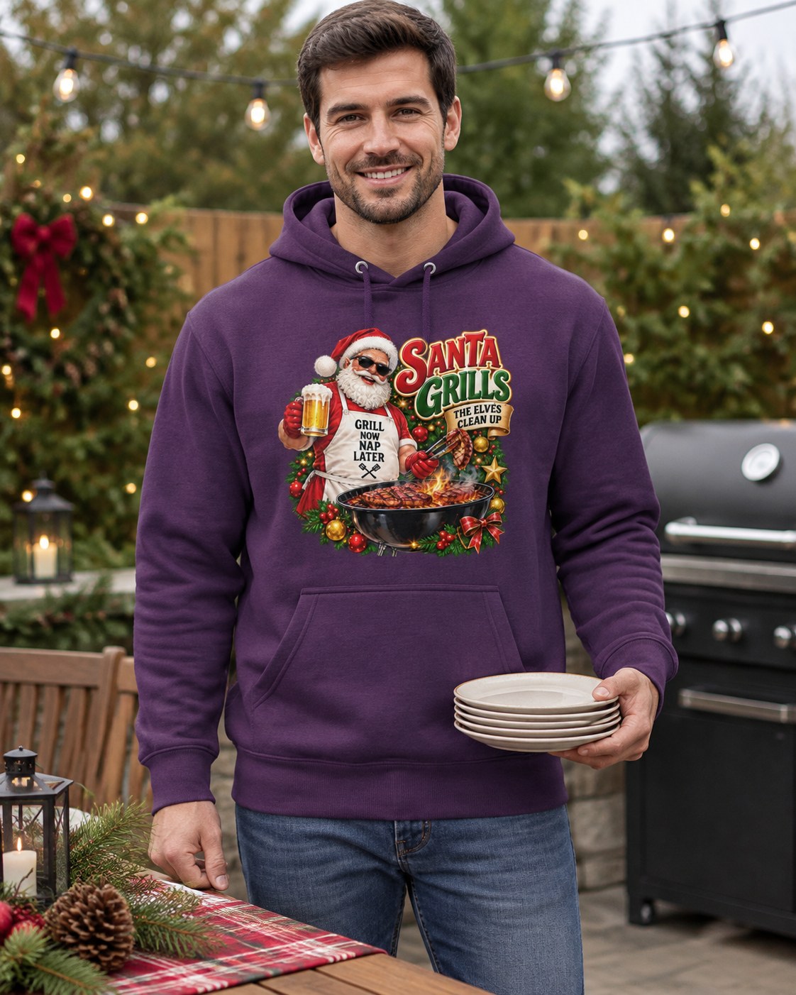Man wearing a purple Santa grilling Christmas hoodie, holding plates beside a barbecue and Christmas-decorated table