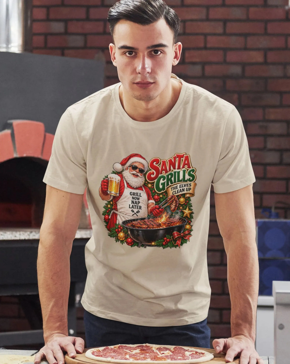 Man wearing a beige Santa grilling Christmas shirt, leaning over a counter with an uncooked pizza in front of a brick pizza oven.