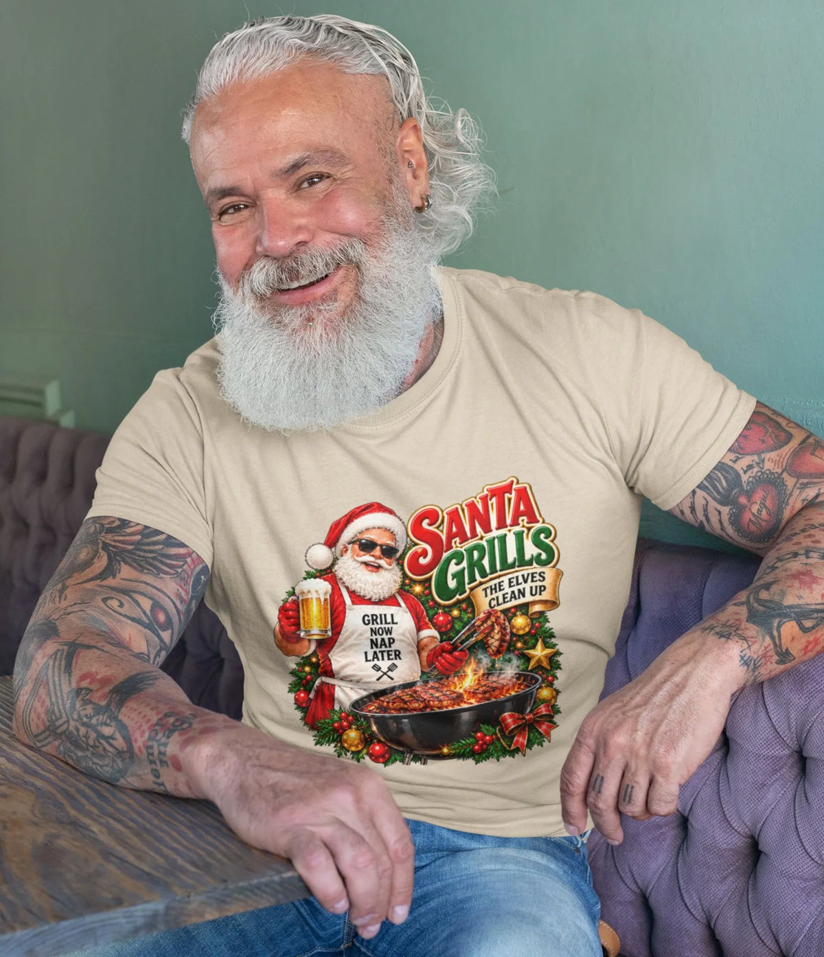 Smiling, white-bearded man wearing a beige Santa grilling Christmas shirt and blue jeans, seated beside a wooden table with the festive BBQ graphic visible.