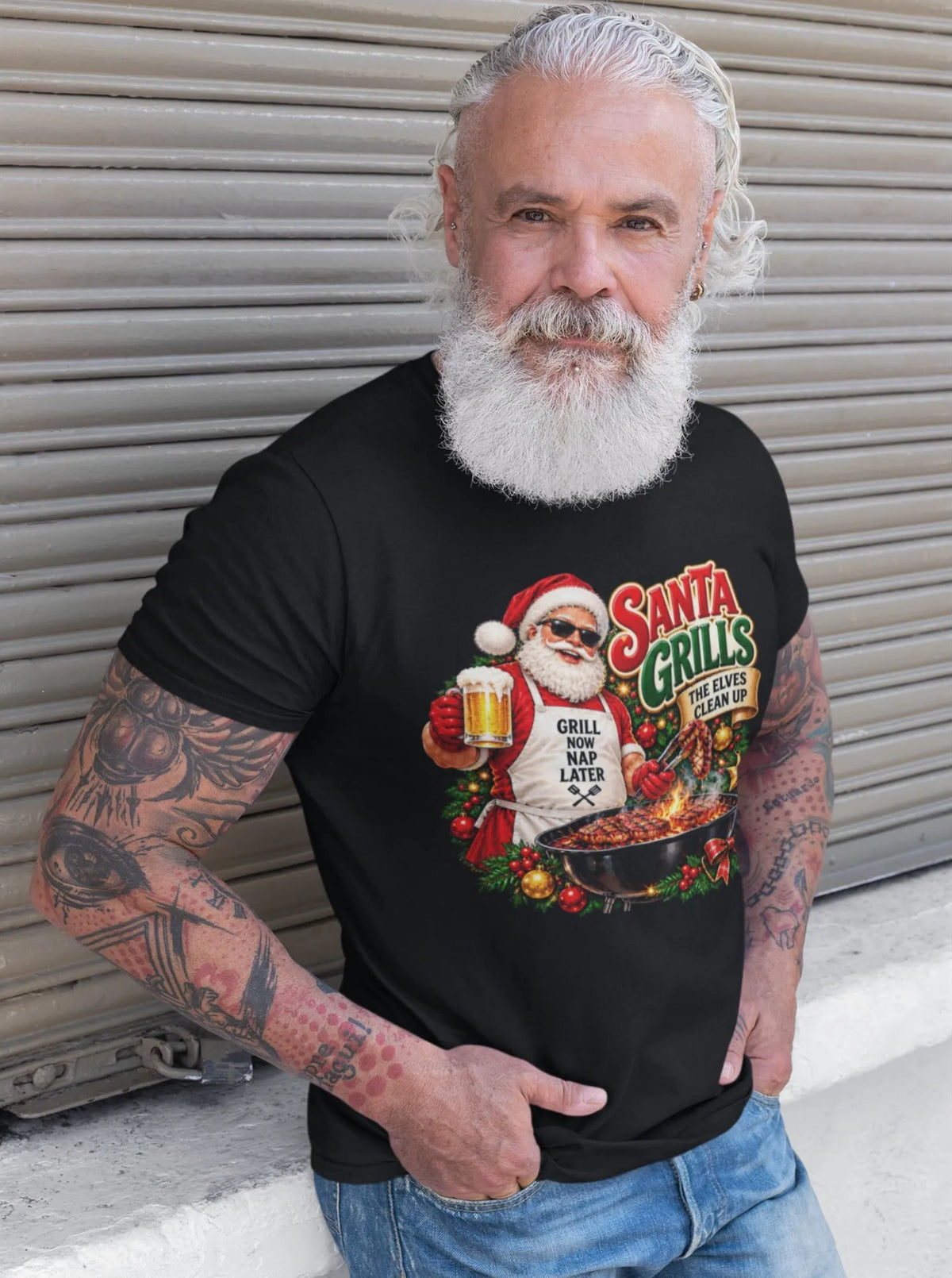 White-bearded man with tattooed arms wearing a black Santa grilling Christmas shirt and blue jeans, standing beside a metal shutter with the BBQ Santa graphic visible