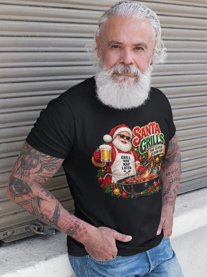 White-bearded man with tattooed arms wearing a black Santa grilling Christmas shirt and blue jeans, standing beside a metal shutter with the BBQ Santa graphic visible