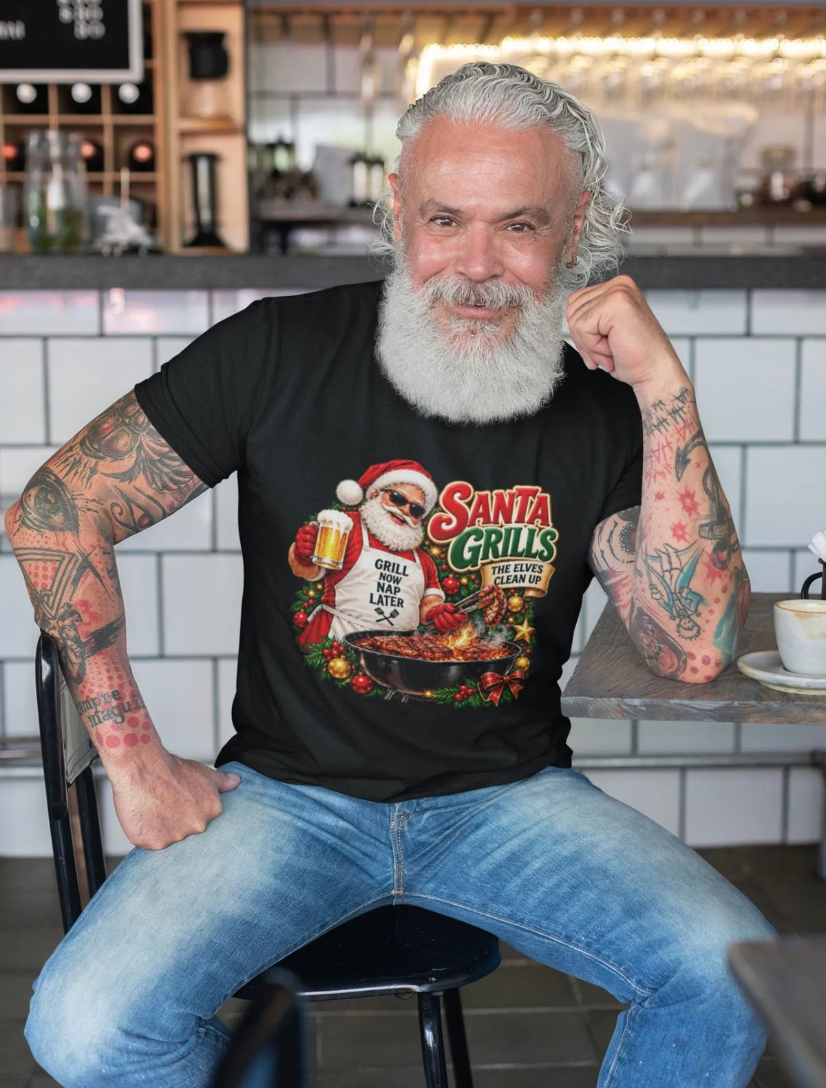 Smiling, white-bearded man wearing a black Santa grilling Christmas shirt and blue jeans, seated at a café table with the festive BBQ graphic visible.