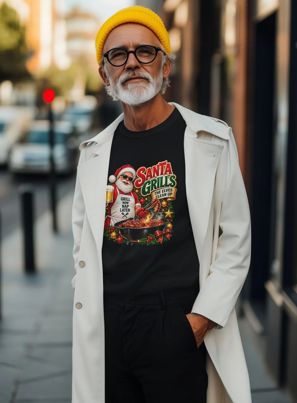 A man wearing a black graphic t-shirt with a 'Santa Grills' Christmas design, styled with a white trench coat, black pants, and a bright yellow beanie, standing on a city street.